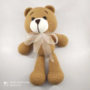 A close-up image of a crocheted amigurumi teddy bear with brown fur and a white bow.