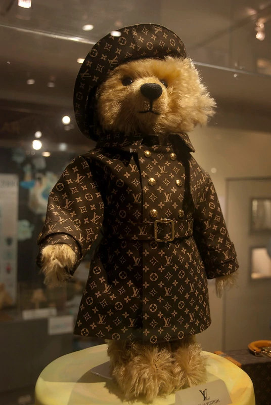 A brown teddy bear wearing a white t-shirt and a brown leather collar with a gold-colored metal tag. The teddy bear is standing on a wooden table with a dark background.