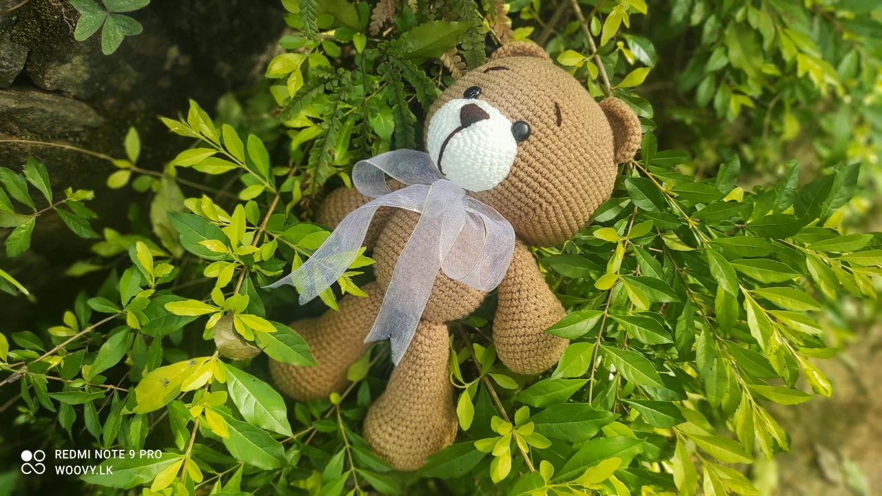 A crocheted teddy bear sitting on a tree stump in a beautiful natural setting.