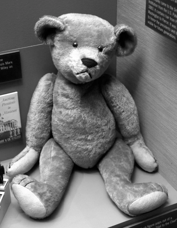 A black and white photograph of a vintage teddy bear believed to have been made by Morris Michtom in the early 1900s. The teddy bear is made of light-colored fur and has black button eyes and a black nose. It is sitting on its hind legs with its front paws stretched out in front of it.