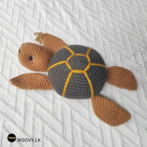 A brown and gray crocheted sea turtle with a yellow stripe around its neck sits on a white bedspread.