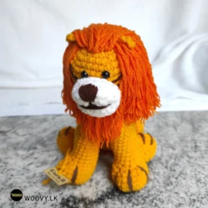 A crocheted lion toy sits on a table. The lion is made of brown and yellow yarn and has a friendly face with a stitched smile.